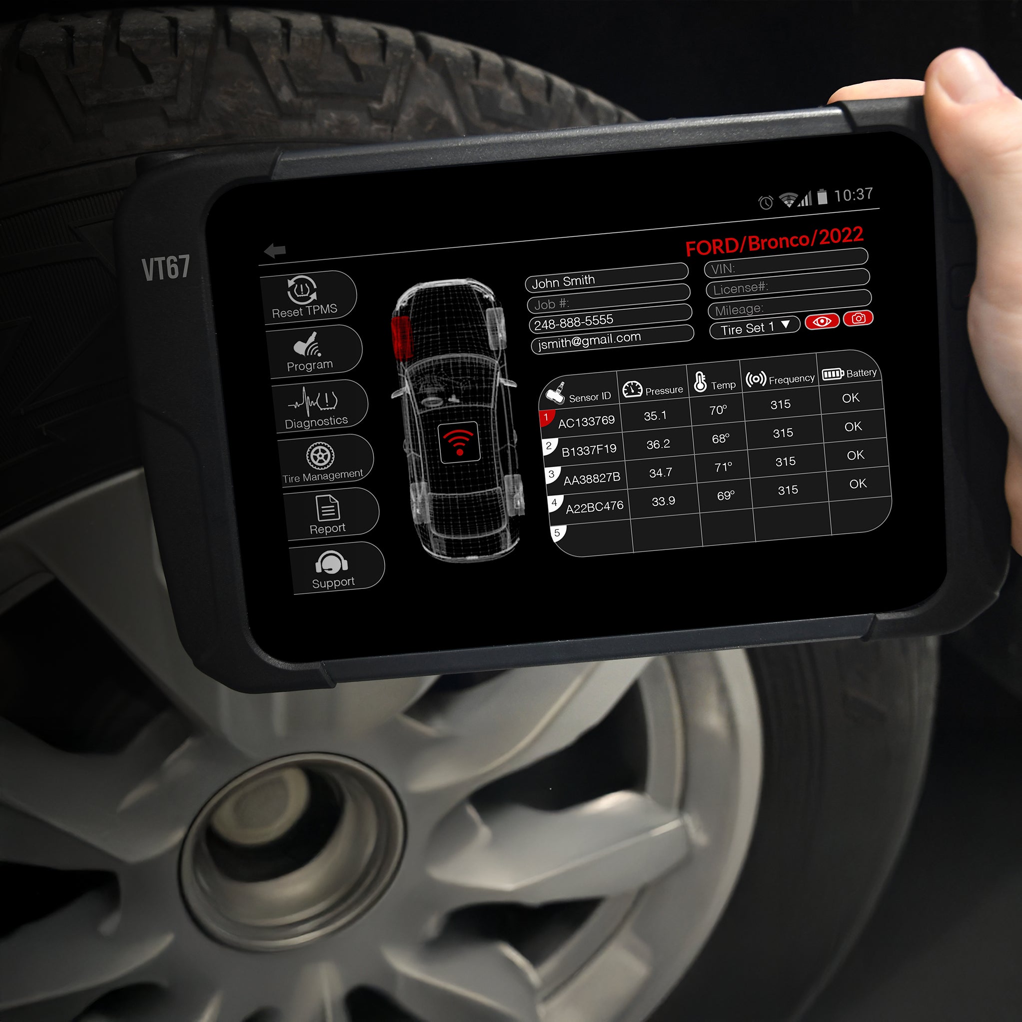 VT67 diagnostic tool held by a person with a car tire in the background
